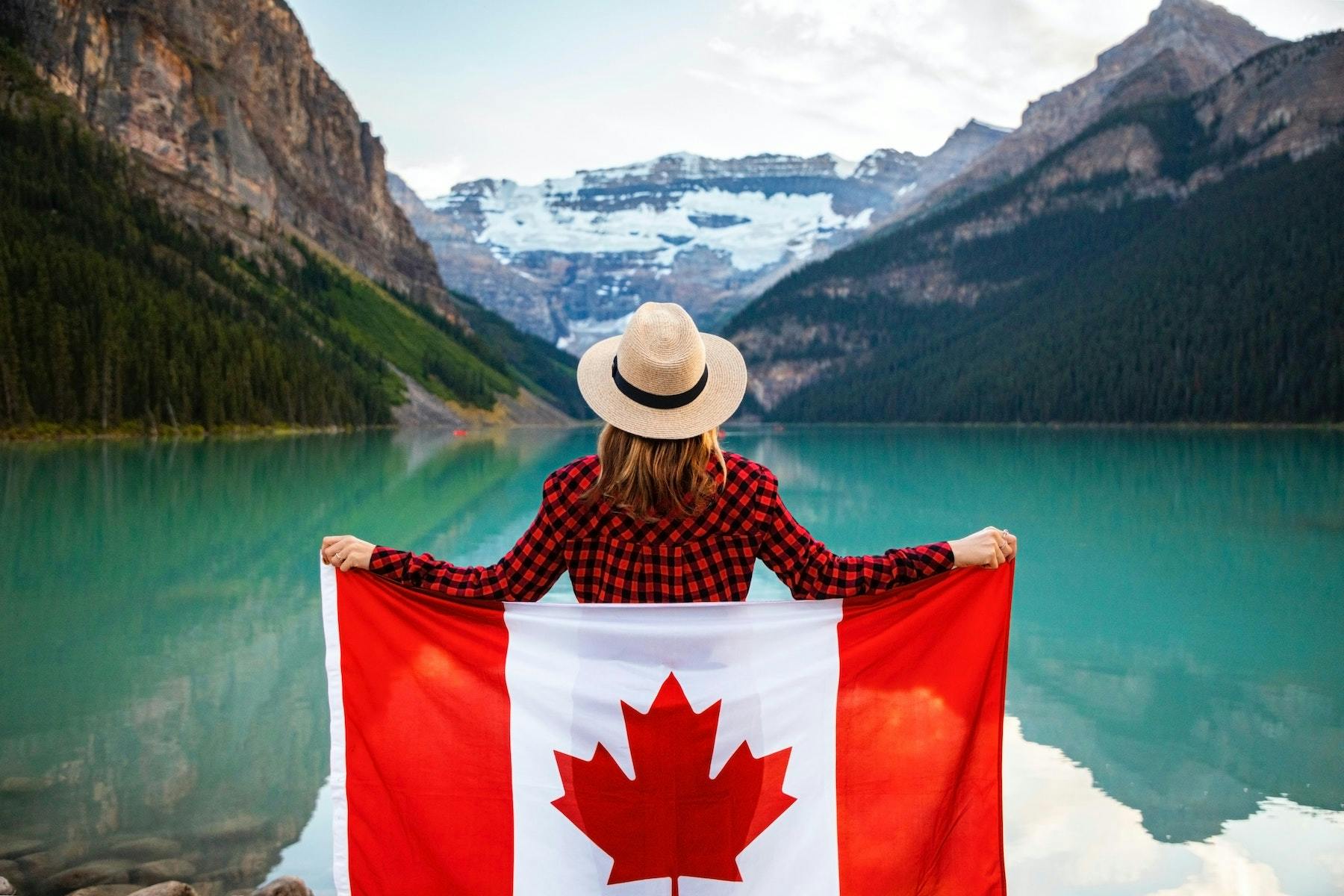 Mulher vestindo camisa xadrez vermelha e preta e chapéu Fedora bege segurando a bandeira do Canadá olhando para um lago